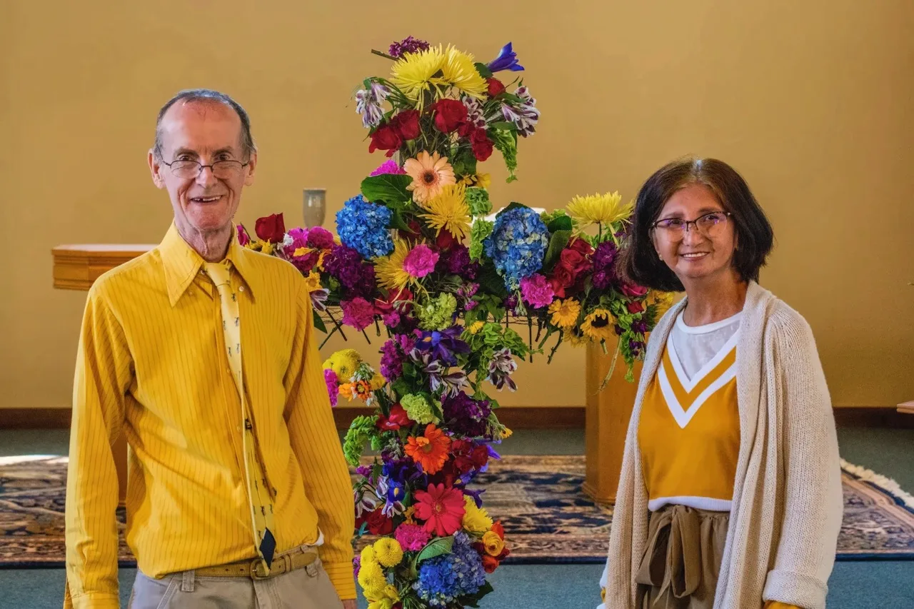 Two church members smiling beside the Easter cross covered in fresh flowers